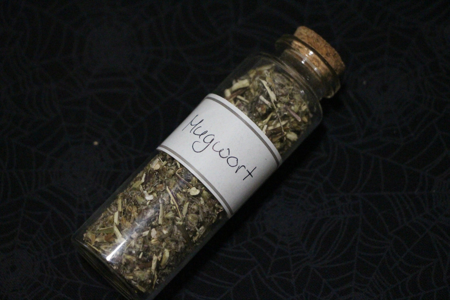 Glass jar of dried mugwort labeled for witchcraft rituals on black spiderweb cloth