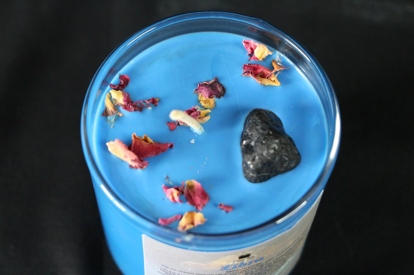 Blue crystal candle with dried flower petals and black stone in glass jar on black background