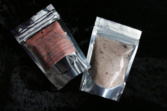 Two silver resealable bags of aromatic pink bath salts with rose petals on black fabric