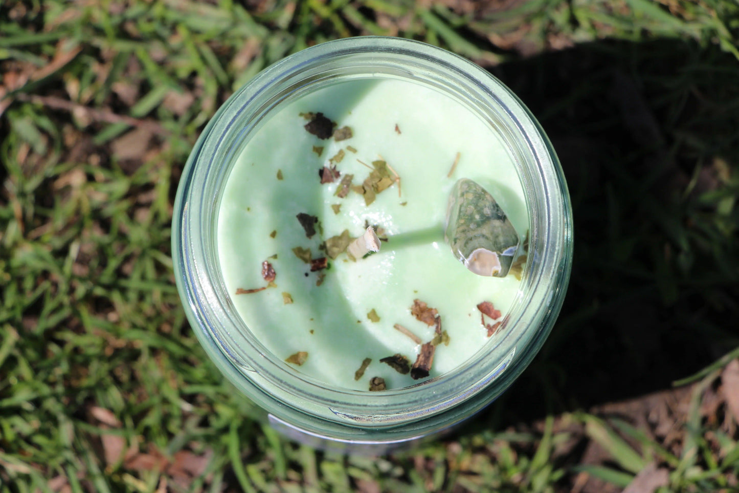 Green crystal candle with herbs in glass jar, photographed outdoors on grass