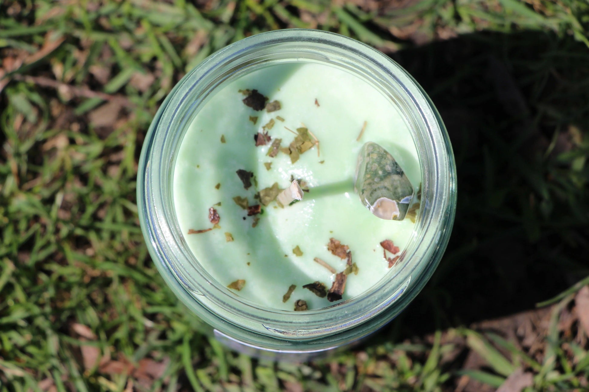 Green crystal candle with herbs in glass jar, photographed outdoors on grass