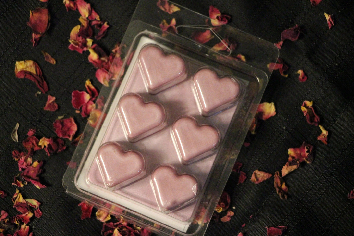 Pink heart-shaped wax melts in clamshell packaging on black cloth with dried rose petals