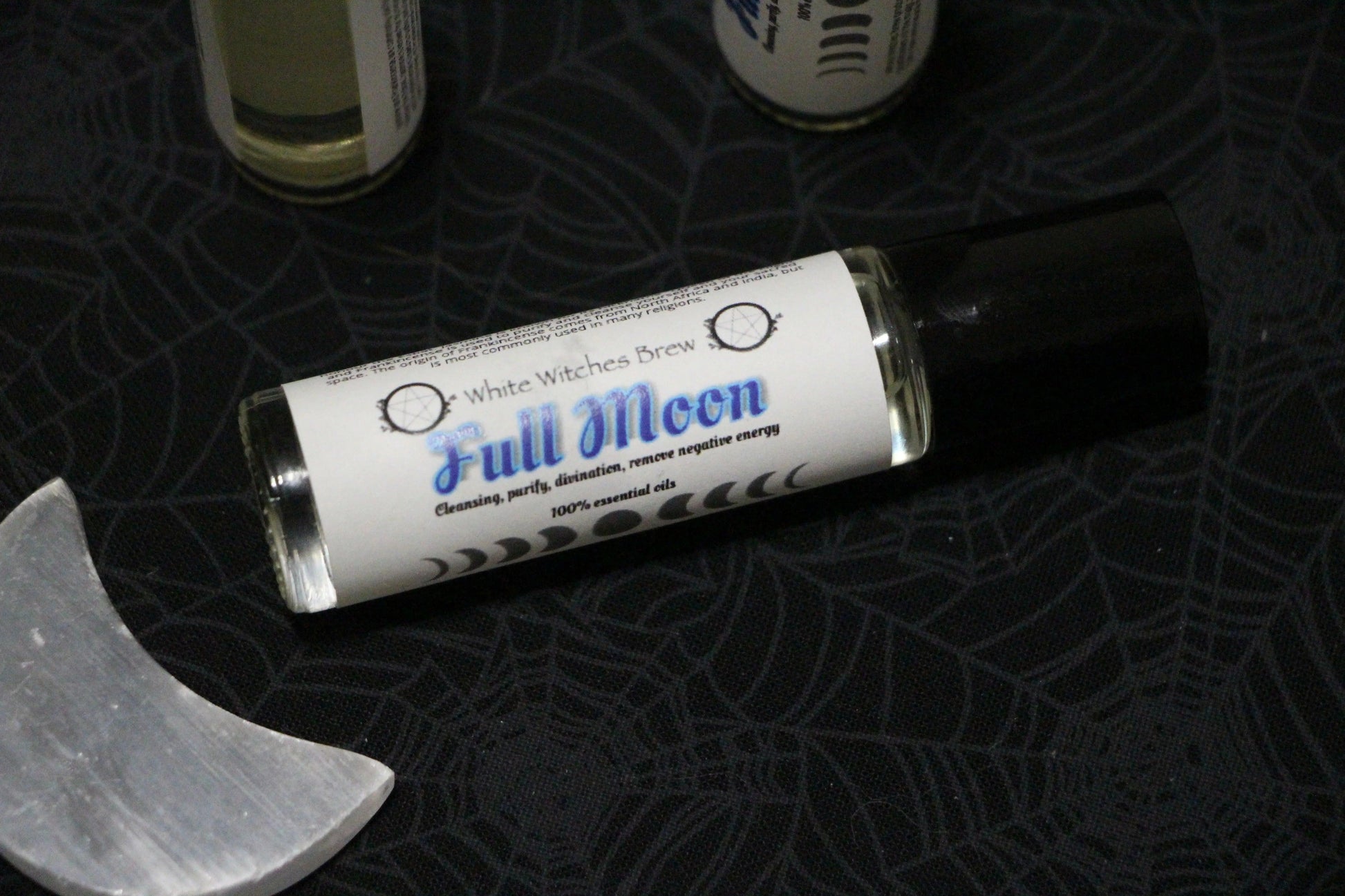 White Witches Brew Full Moon essential oil roller on spiderweb fabric with a crescent-shaped crystal