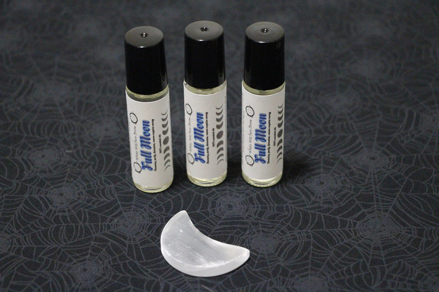 Three Full Moon essential oil roller bottles with black caps and a selenite crescent on spider web fabric