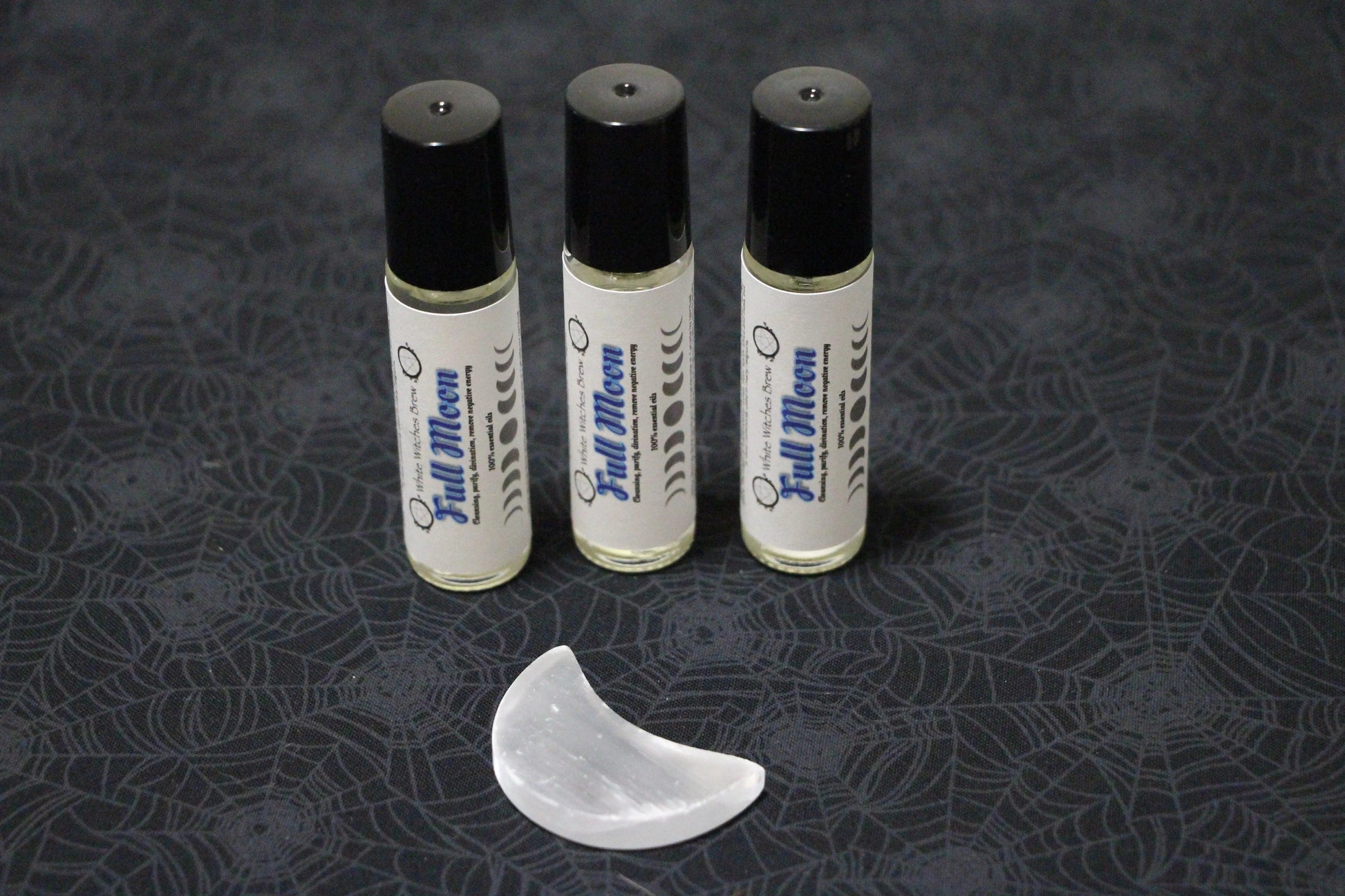 Three Full Moon essential oil roller bottles with black caps and a selenite crescent on spider web fabric
