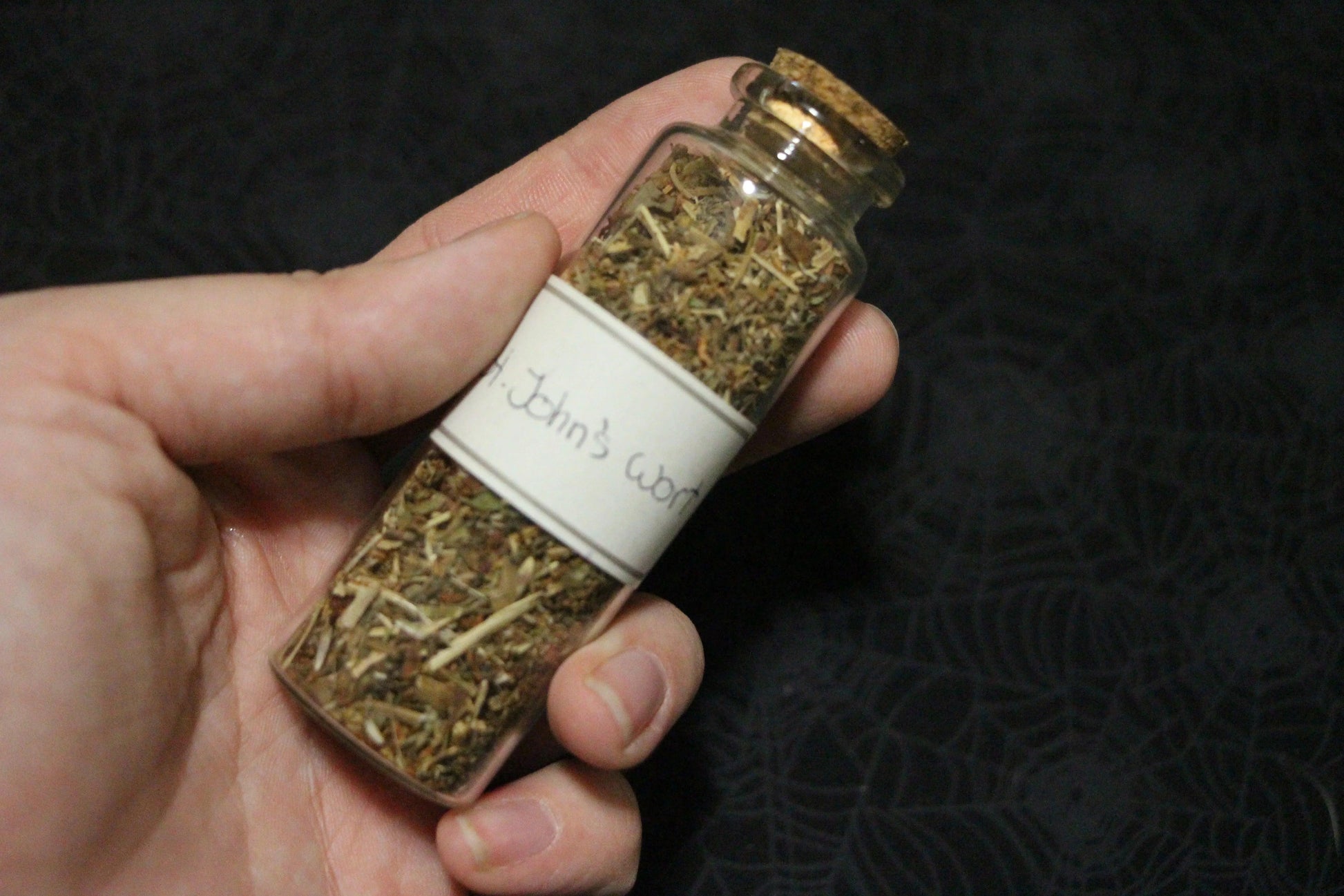 Hand holding a glass jar of dried St. John's wort herbs for witchcraft rituals