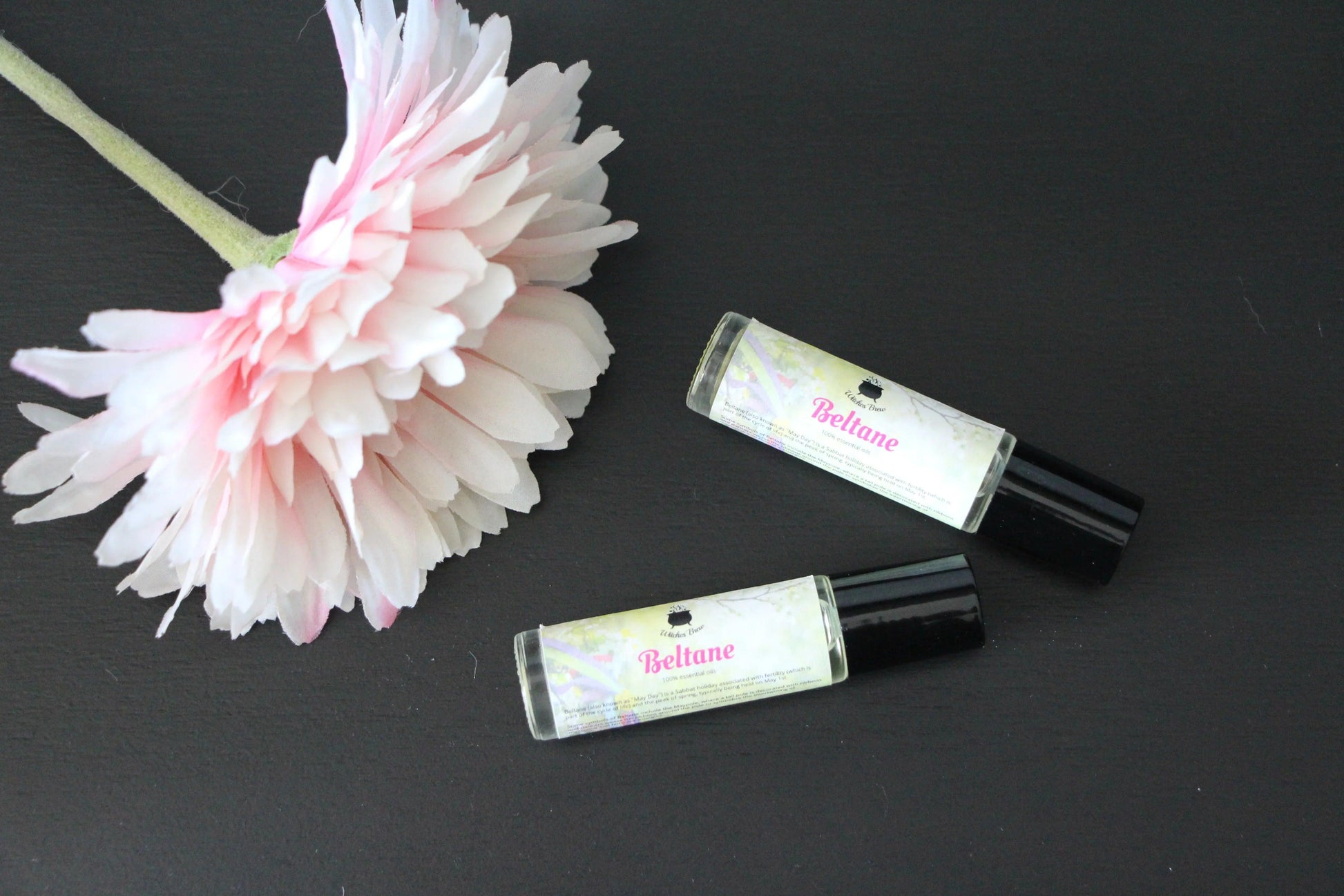Two Beltane essential oil rollers and a pink flower on a black background