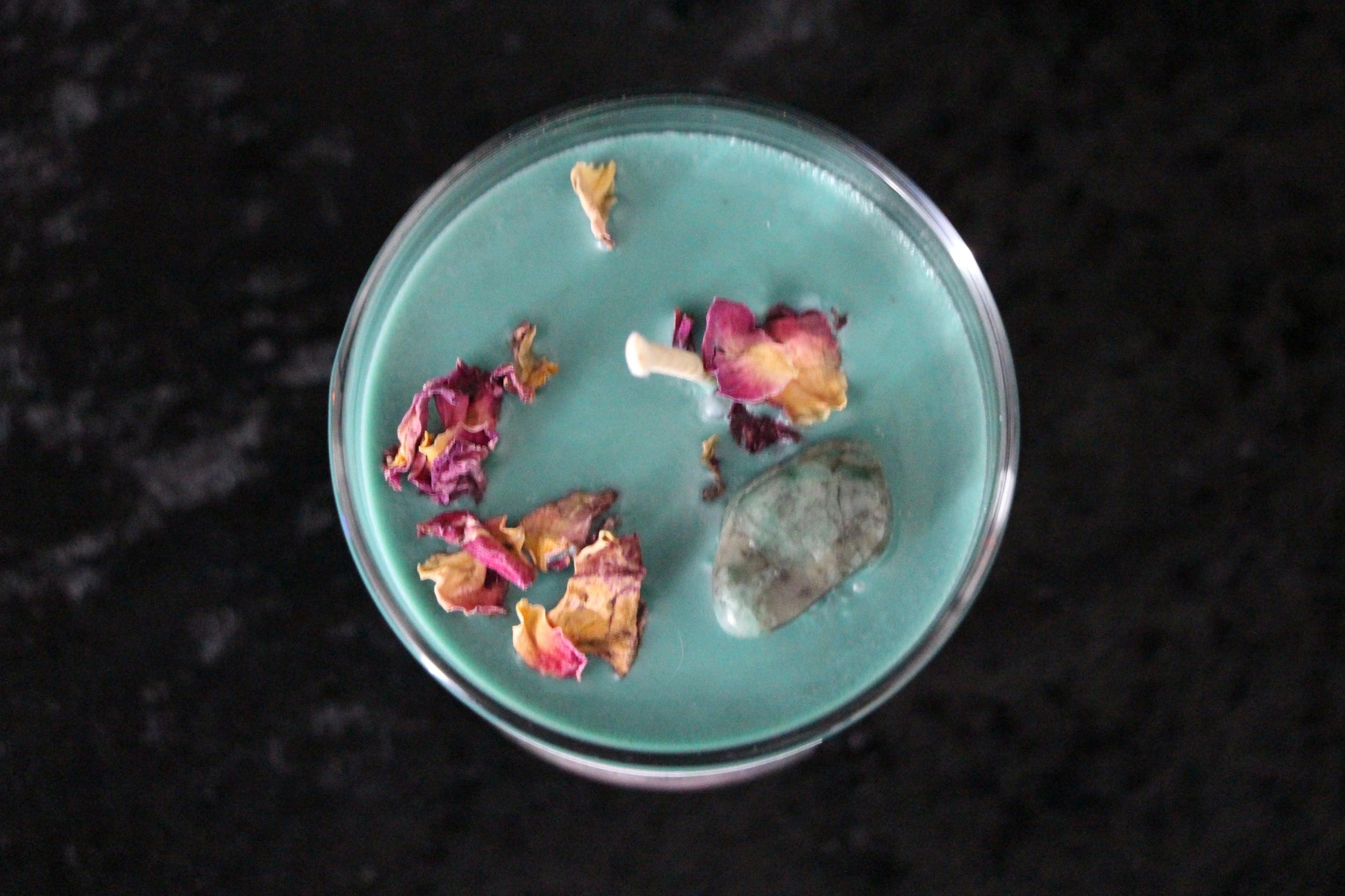 Hand-crafted teal crystal candle with dried rose petals and gemstone on dark background