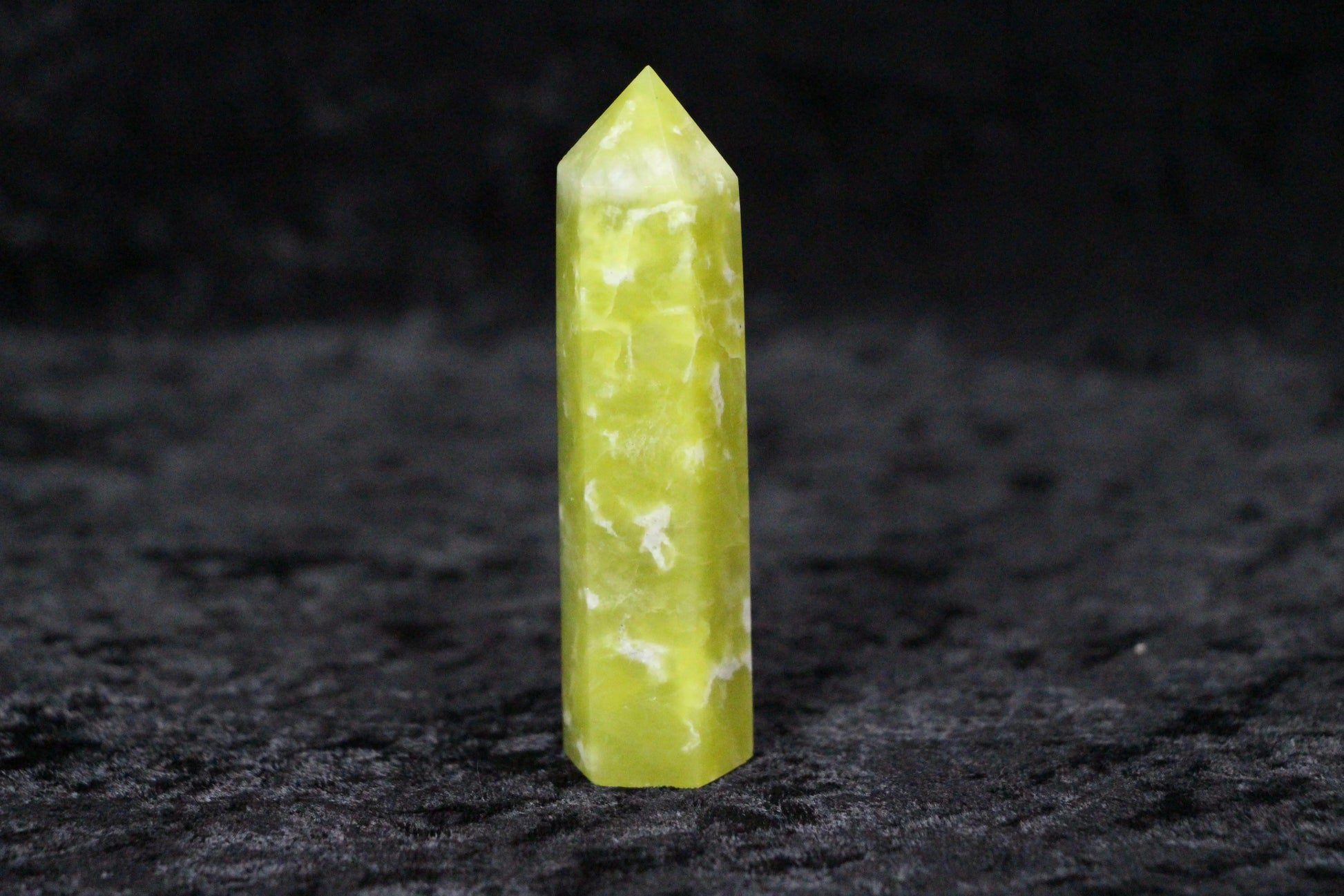 Polished yellow-green serpentine crystal point standing on black textured surface