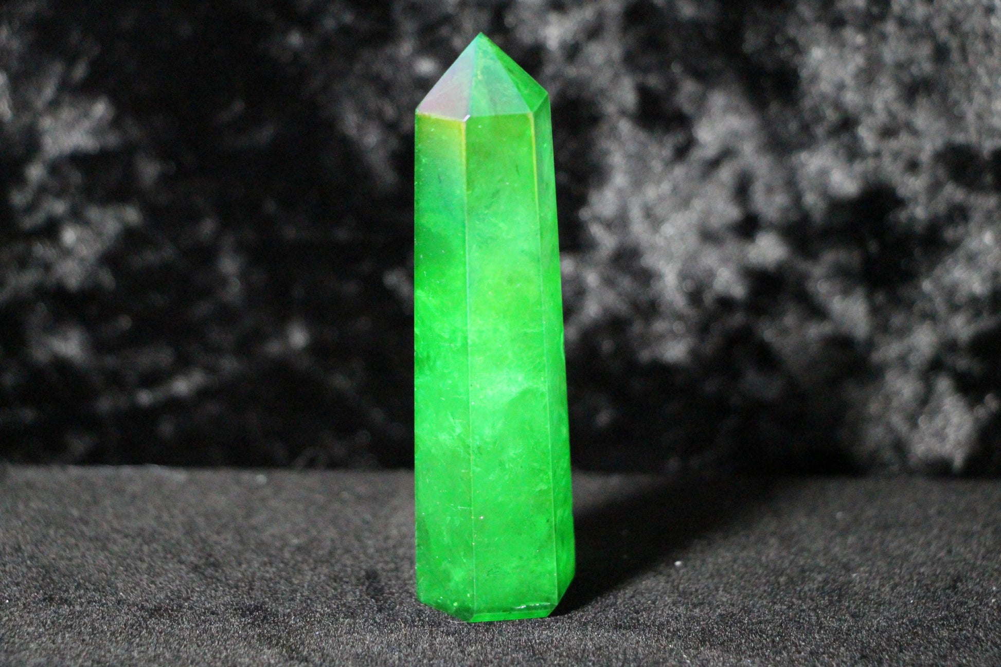 Bright green crystal obelisk standing on a dark textured surface with dark background