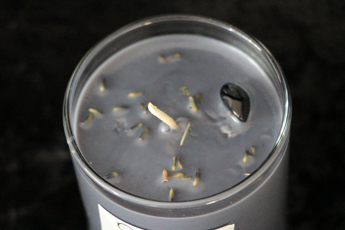 Close-up of a gray scented candle in glass jar with dried herbs and black stone on surface