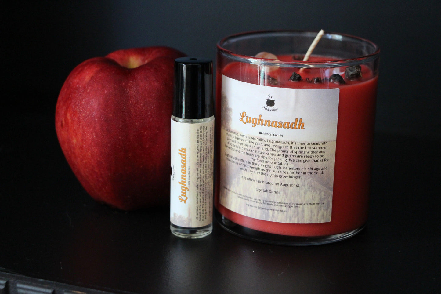 Red apple, Lughnasadh essential oil roller, and red crystal candle by Witches' Brew on black background