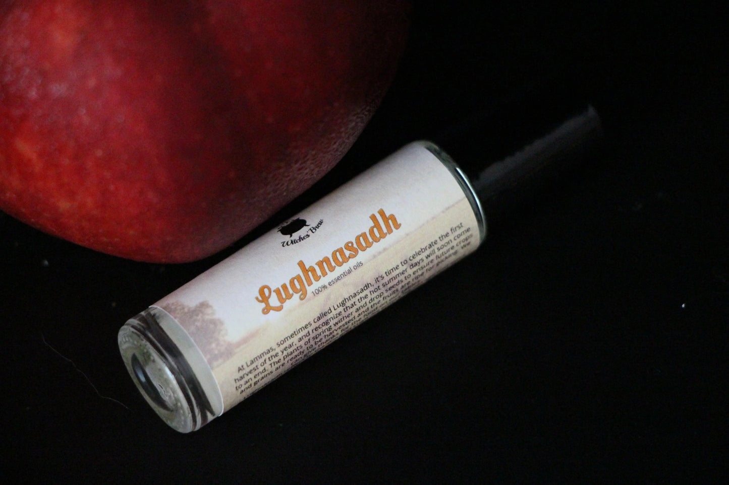 Lughnasadh essential oil roll-on by Witches' Brew beside a red fruit on black background