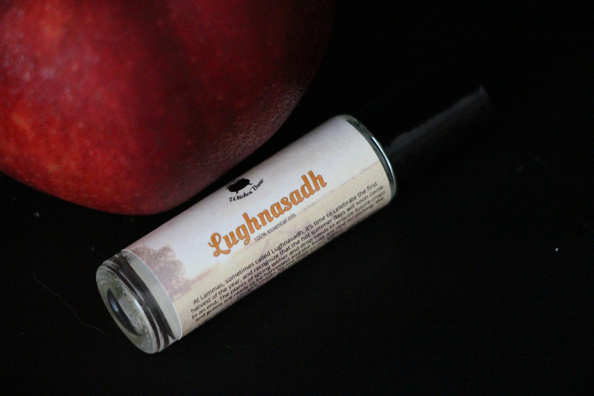 Lughnasadh essential oil roll-on by Witches' Brew beside a red fruit on black background