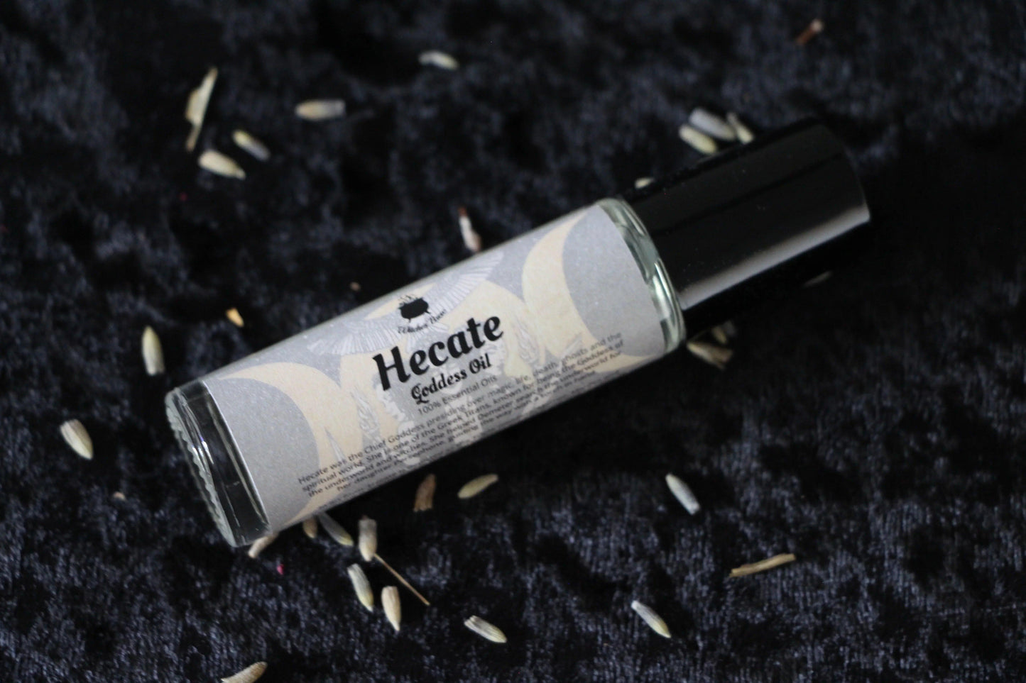 Hecate Goddess Oil essential oil roller bottle on black textured fabric with scattered dried herbs