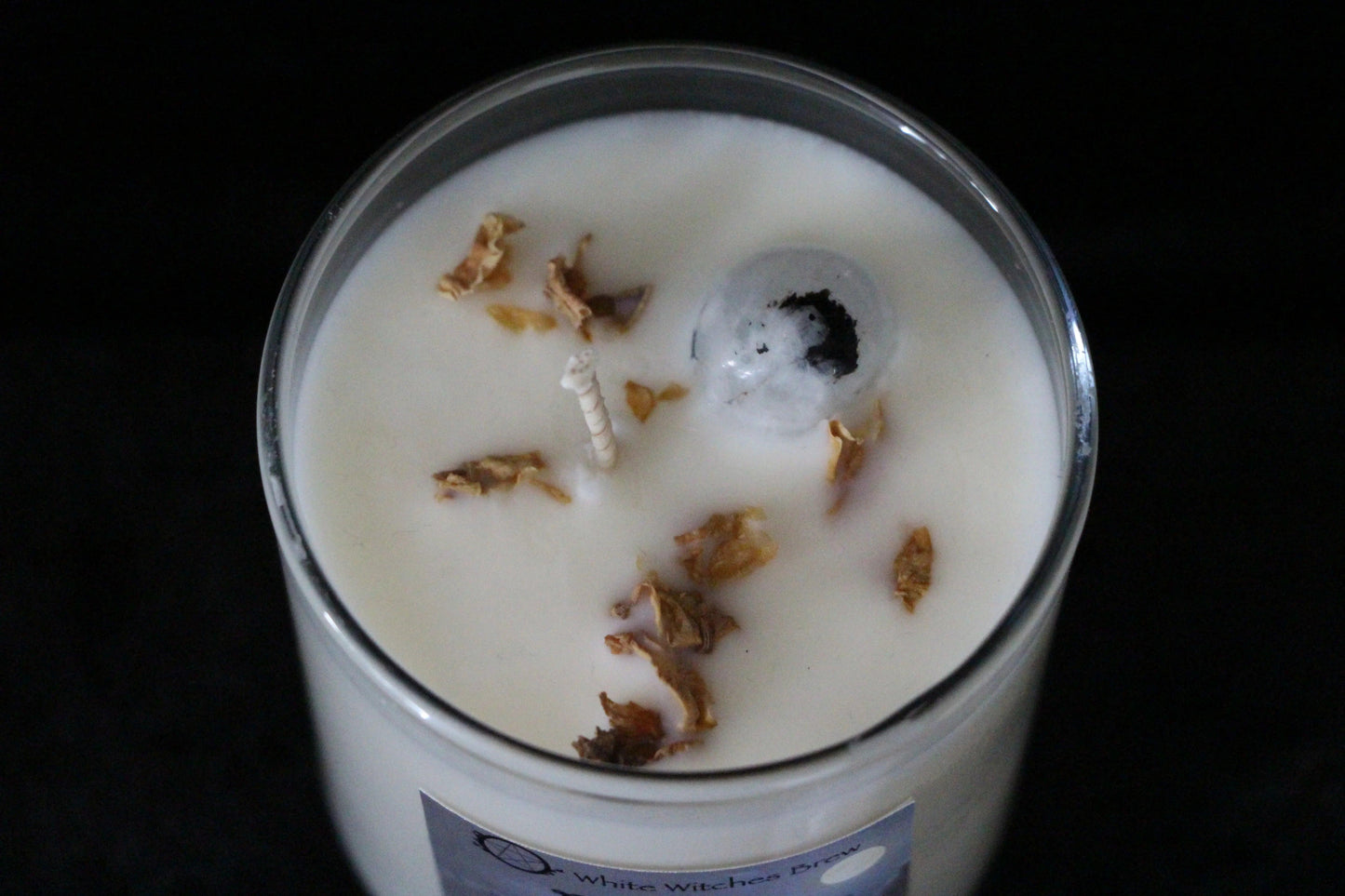White wax candle in glass jar with dry flower petals and a clear crystal, labeled White Witches Brew