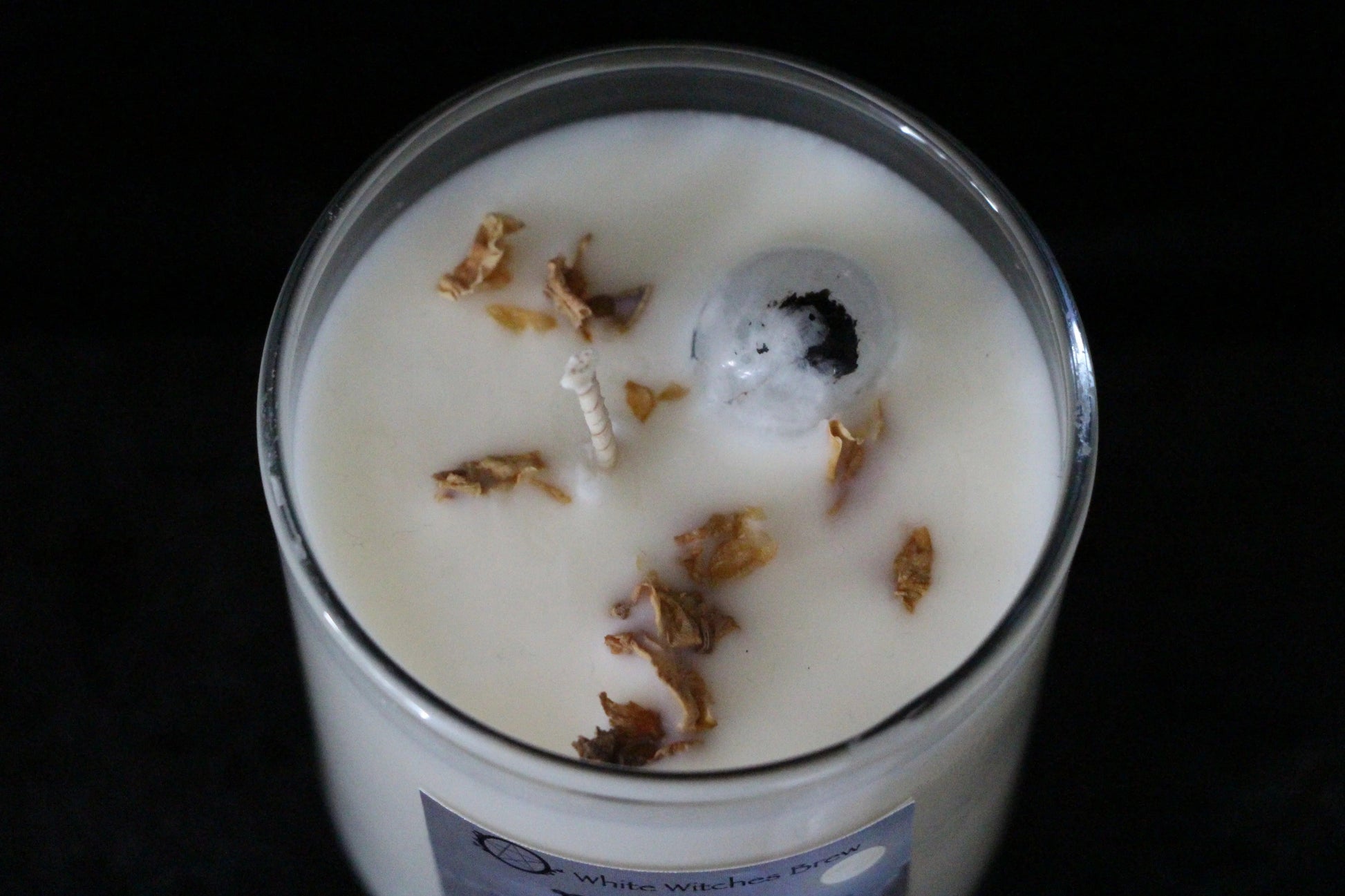 White wax candle in glass jar with dry flower petals and a clear crystal, labeled White Witches Brew