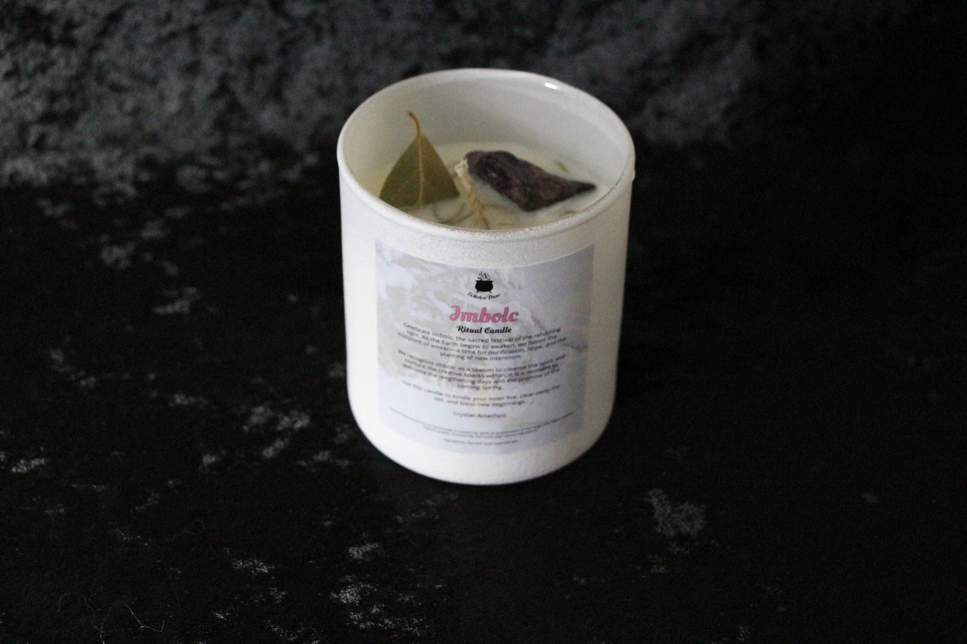 Imbolc ritual candle with herbs and crystal in white container on dark textured surface