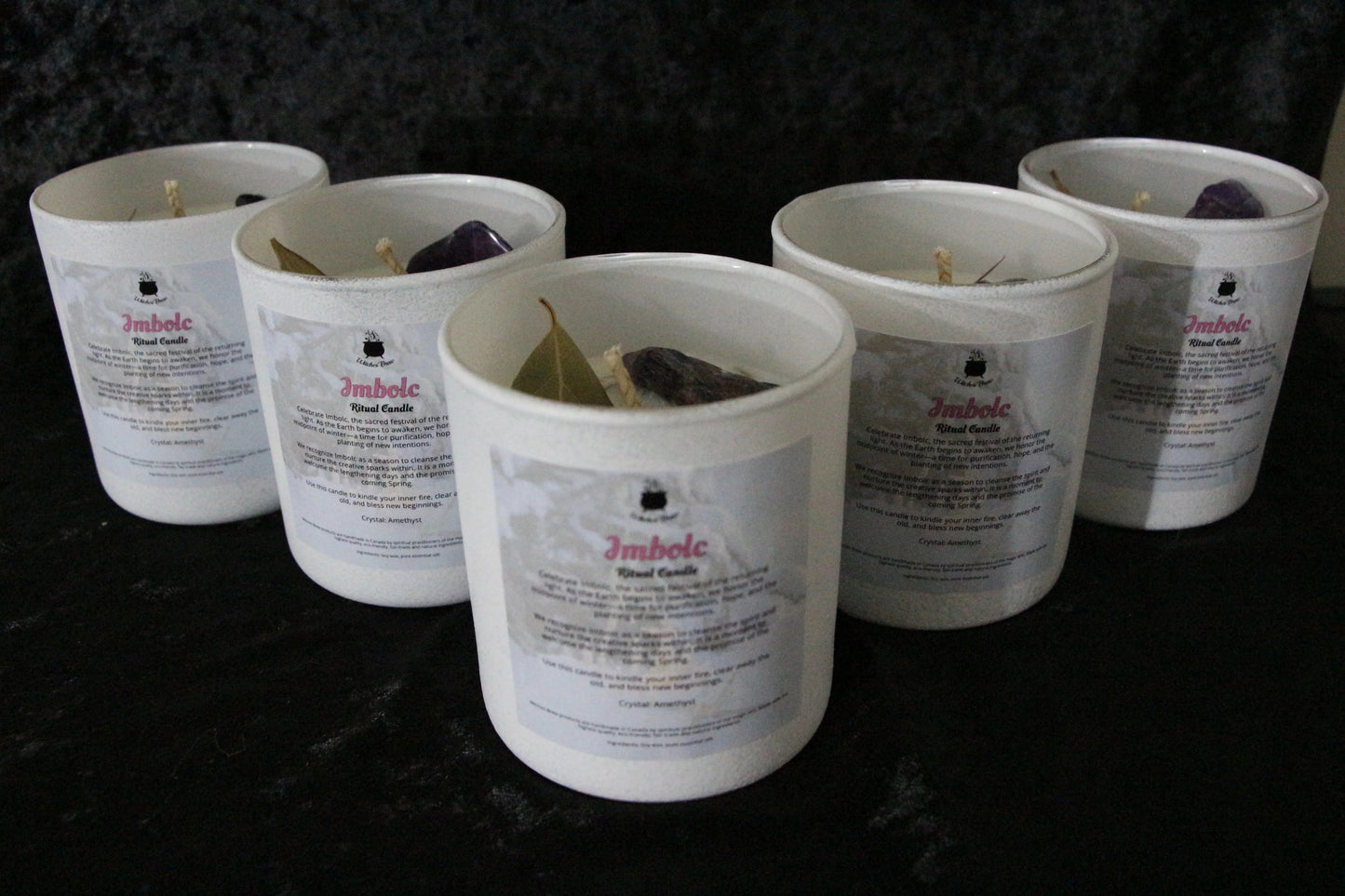 Five white Imbolc ritual candles with herbs and crystals on a dark fabric background